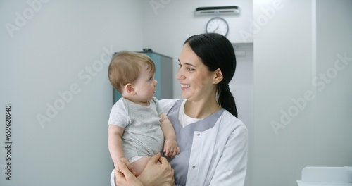Female pediatrician stands in middle of bright hospital room and looks at camera. Adult doctors holds baby. Little boy looks at camera and holds hand of medic. Medical staff at work in modern clinic.