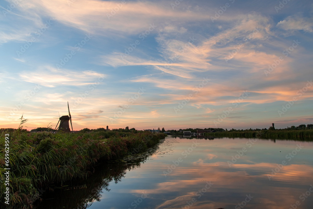 Sunset view over tranquil water with reed beds of a canal with perfect reflection of sunset clouds in water next to silhouette of iconic 18th-century windmill in Kinderdijk, the Netherlands Sunset view over tranquil water with reed beds of a canal with perfect reflection of sunset clouds in water next to silhouette of iconic 18th-century windmill in Kinderdijk, the Netherlands