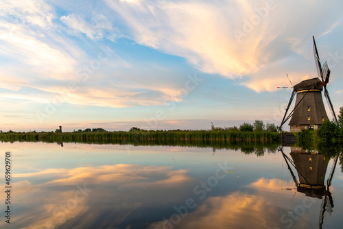 Typical Dutch windmill along long row of reed beds in the Kinderdijk area, the Netherlands with a near perfect reflection of the windmill, reed beds and clouds in the tranquil water during sunset photo