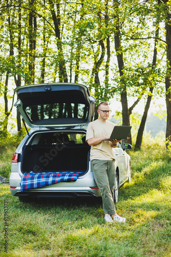 Cheerful traveler, young man sitting in open trunk of car, working with laptop, outdoors, concept of business, travel, vacation © Дмитрий Ткачук