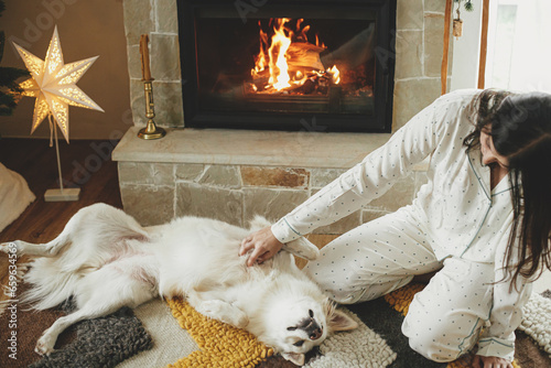 Cozy christmas morning. Beautiful woman in stylish pajamas relaxing and playing with cute white dog at warm fireplace in festive decorated living room. Merry Christmas! Winter holidays with pet
