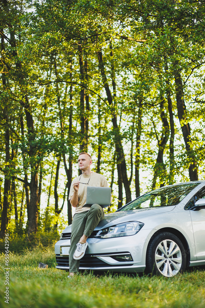 A serious man in glasses is working on a laptop on the hood of his car. Remote work in nature. Work on a laptop online. Working on a computer while traveling. A serious man in glasses is working on a laptop on the hood of his car. Remote work in nature. Work on a laptop online. Working on a computer while traveling.