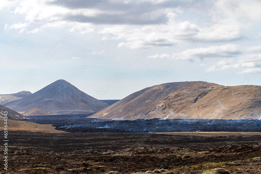 Panoramic view over landscape and smoking lava field near the fissure of Litli-Hrútur during the 2023 eruption near the mountain Fagradalsfjall, Iceland against a white clouded blue sky
