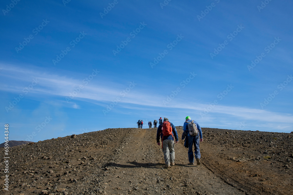 Low angle view of hikers on a steep path in volcanic landscape on Reykjanes Peninsula, Iceland, near sites of 2021-2022 eruptions near mountain Fagradalsfjall volcano area against blue sky