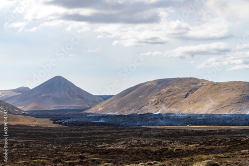 Panoramic view over landscape and smoking lava field near the fissure of Litli-Hrútur during the 2023 eruption near the mountain Fagradalsfjall, Iceland against a white clouded blue sky