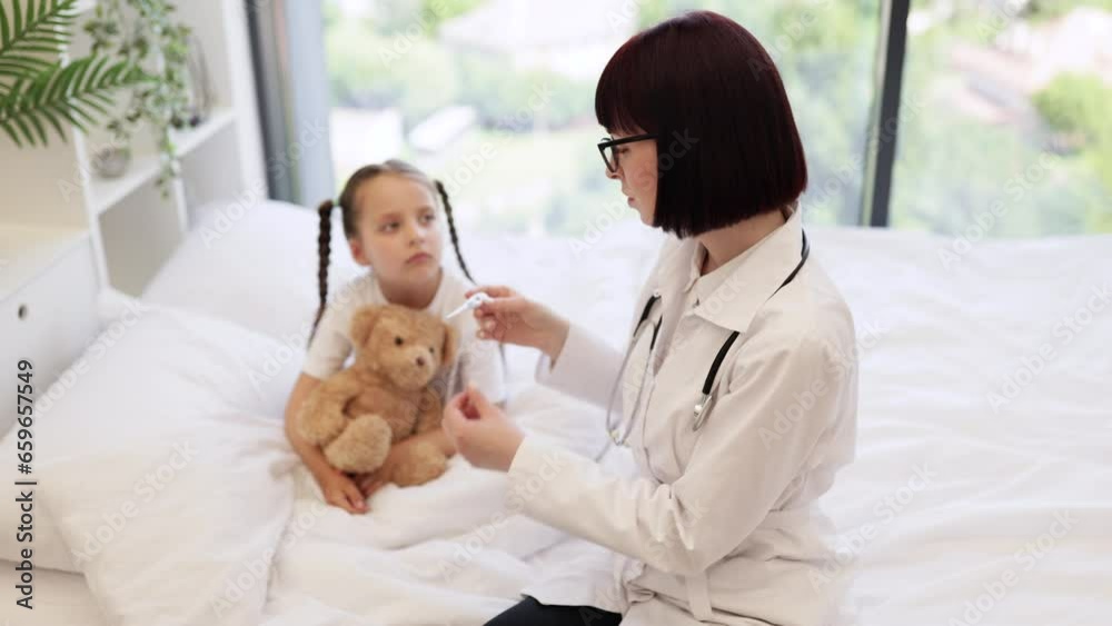 Young girl patient sitting in white bed under watchful care of doctor conducting thorough examination and checking temperature. Cute child hugging toy and preparing to taking medicine. Young girl patient sitting in white bed under watchful care of doctor conducting thorough examination and checking temperature. Cute child hugging toy and preparing to taking medicine.
