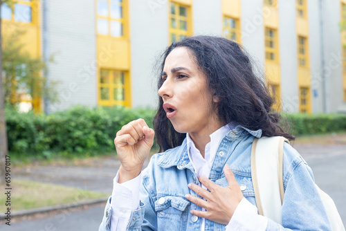 Woman on the street on the background a hospital or educational institution experiences a coughing attack. Girl coughs into her fist with her other hand, holds her chest, breathes heavily, gets sick.