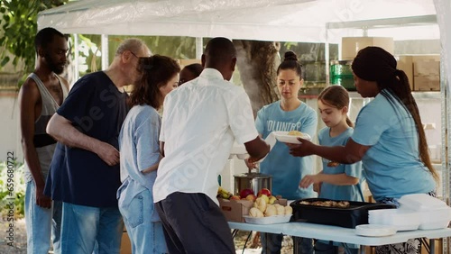 At homeless shelter team of voluntary people dedicatedly packaging food parcels to give to less fortunate. Poor and needy individuals patiently waiting to be served free meal by charity workers.