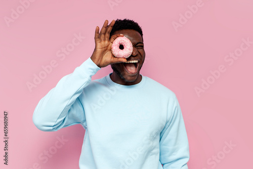 shocked african american man in blue sweater holding sweet donuts on pink isolated background