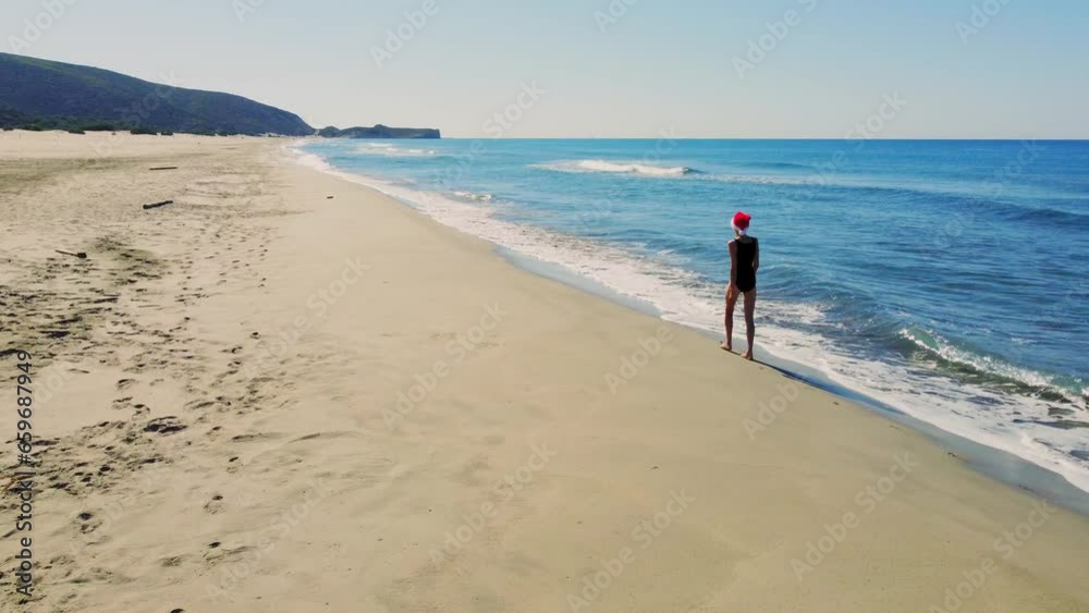 Young beautiful slender woman in Santa Claus hat walking on sandy beach, rejoicing in Christmas or New Year. Shooting with drone. Weekend Sea or Ocean. Resort Vacation. Young beautiful slender woman in Santa Claus hat walking on sandy beach, rejoicing in Christmas or New Year. Shooting with drone. Weekend Sea or Ocean. Resort Vacation.