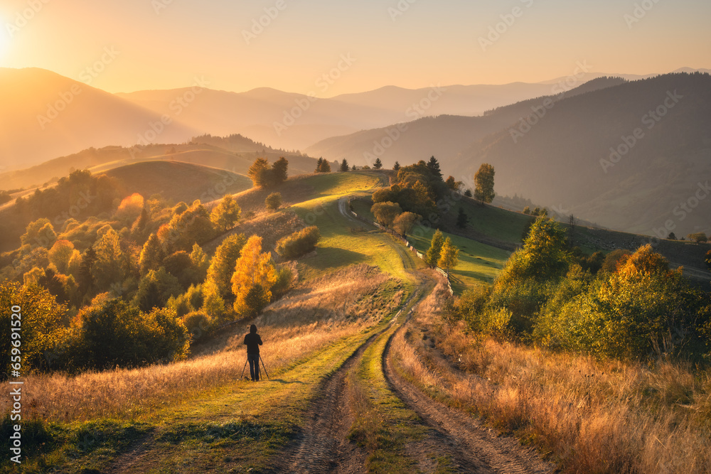 Carpathian mountain valley with beautiful hills in haze, man, orange trees at sunset in autumn in Ukraine. Colorful landscape with dirt road, meadows, forest, grass, golden sunlight in fall. Nature - obrazy, fototapety, plakaty 