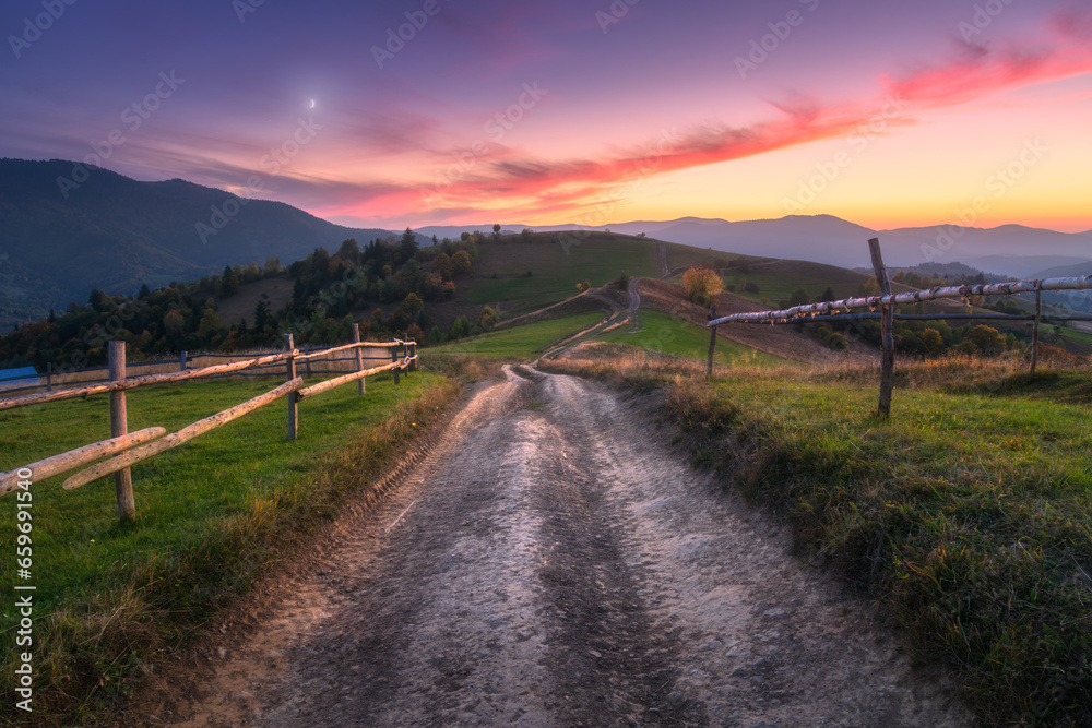 Rural dirt road, wooden fence, green hills in mountain valley at sunset in autumn. Landscape with country road, meadows, trees, purple sky with pink clouds at twilight. Carpathians, Ukraine. Nature