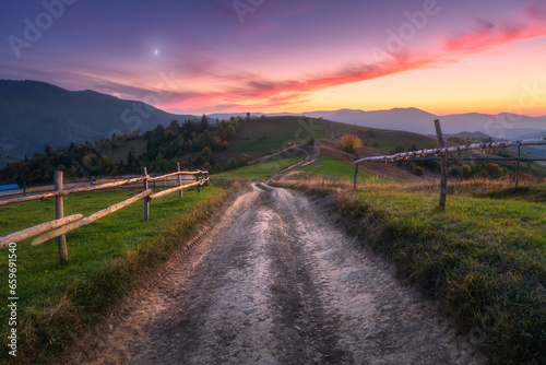 Rural dirt road, wooden fence, green hills in mountain valley at sunset in autumn. Landscape with country road, meadows, trees, purple sky with pink clouds at twilight. Carpathians, Ukraine. Nature