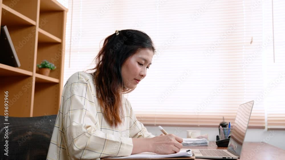 Young Asian woman, office worker, is sitting working in office, busy with work on laptop computer, signing documents that office brought for you sign, It is documented work guideline. Young Asian woman, office worker, is sitting working in office, busy with work on laptop computer, signing documents that office brought for you sign, It is documented work guideline.