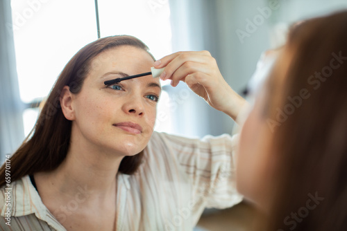 Attractive young woman putting on black mascara in front of a mirror in the bedroom