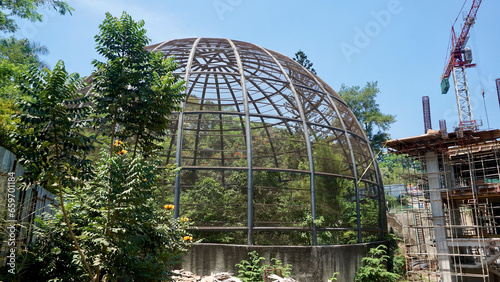 Large metal dome aviary for bird conservation next to a building construction site where crane stand there © thebaikers