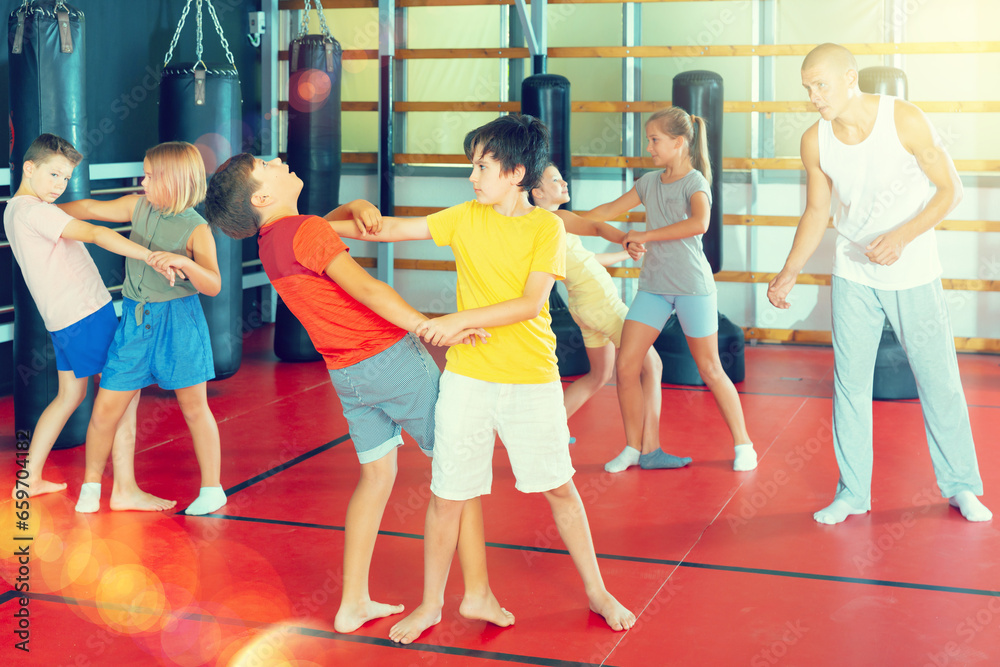 Fototapeta premium Positive kids in pair exercising self-defense movements during group class with male coach