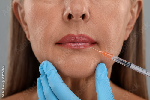 Doctor giving lips injection to senior woman on light grey background  closeup. Cosmetic surgery