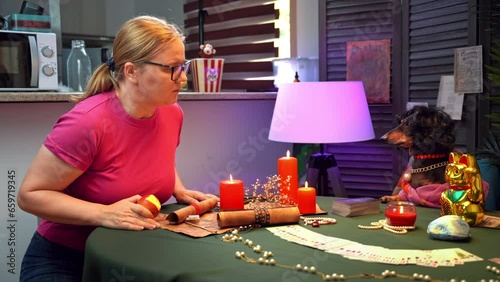 Naive woman with glasses sits down at fortune teller table, shakes head in resignation. Dachshund dog in wig, fortune teller costume sits in esoteric atmosphere with candle, fortune telling tarot card photo