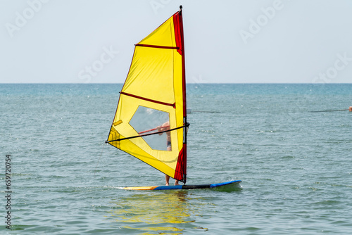 A man rides a blue board with a yellow sail in sunny weather at a tropical resort. A weak wind does not inflate the sail well.