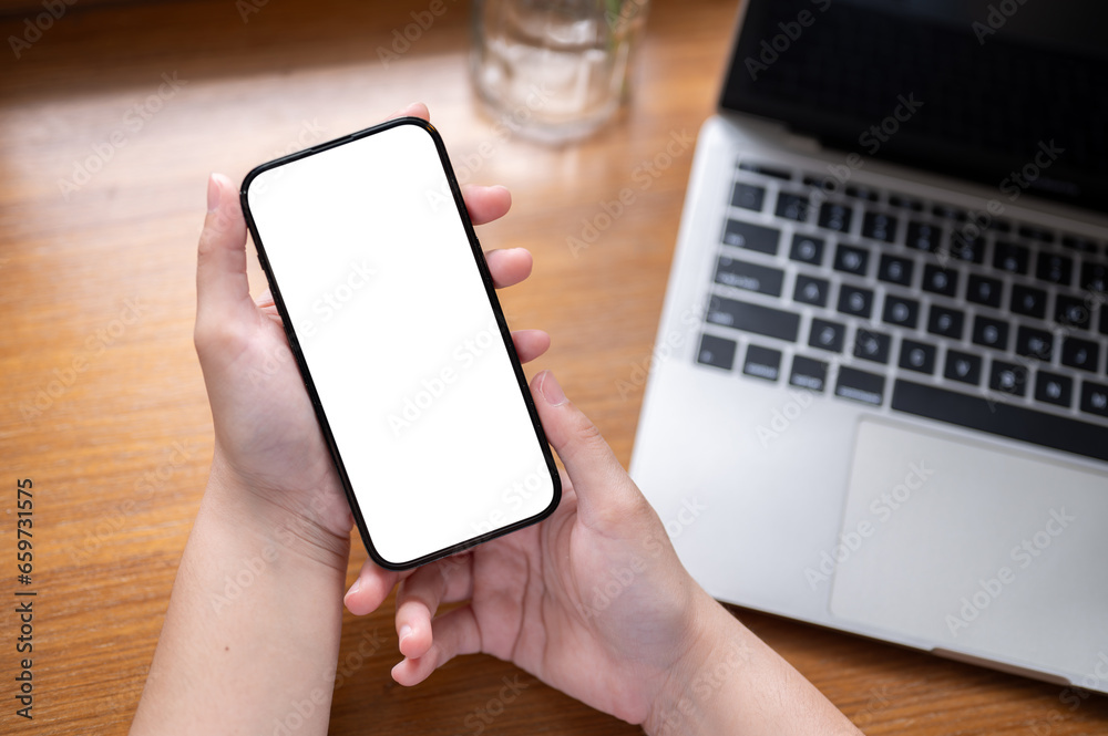 Close-up image of a white-screen smartphone in a woman's hands above a wooden desk with a laptop. Close-up image of a white-screen smartphone in a woman's hands above a wooden desk with a laptop.