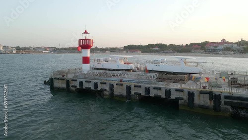 Aerial shot drone pans left along pier with red and white lighthouse at sunset