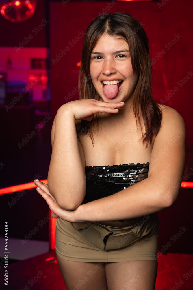 Fototapeta premium young caucasian woman posing from the front and showing her tongue on a background of red lights. night party concept.