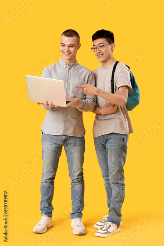 Happy male students with backpack using laptop on yellow background