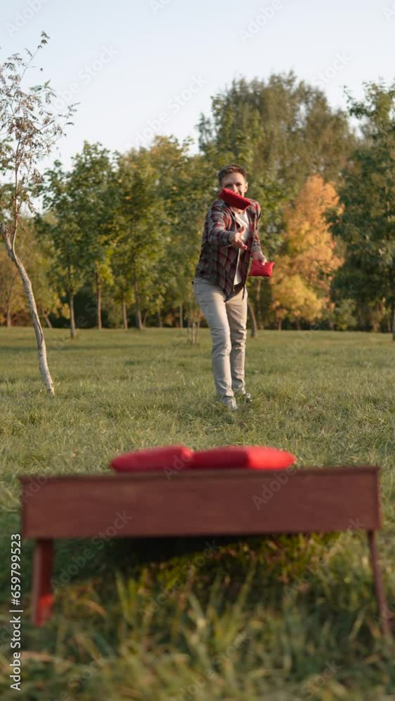 European man toss and hits red bean bag into cornhole. Guy reacts emotionally to successful throw. American wooden game on sunny autumn evening in park. Fun for accuracy on holiday free from work.