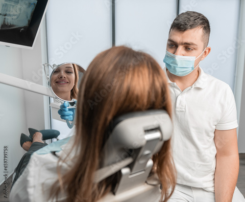 A female patient looks in a mirror showing her a dentist in a clinic. A woman in the clinic receives a full range of dental services  from treatment to teeth alignment.