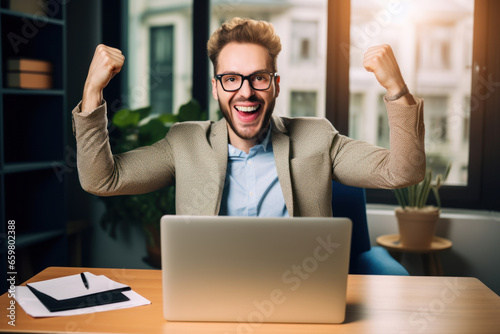 Excited young entrepreneur celebrating success while sitting at workplace with laptop in office