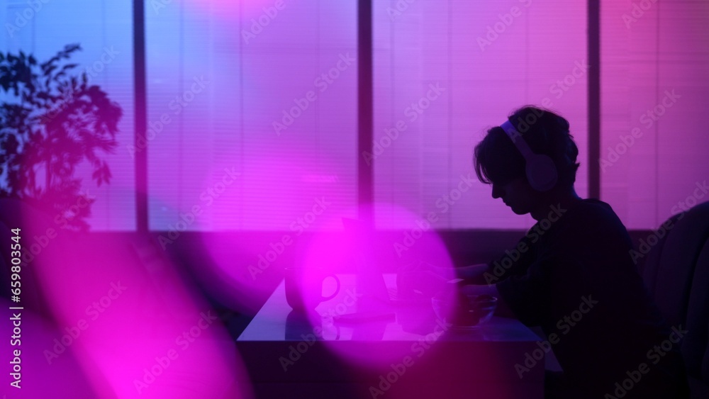 Silhouette of male sitting at the food bar at evening in neon light, wearing headphones, working on laptop.