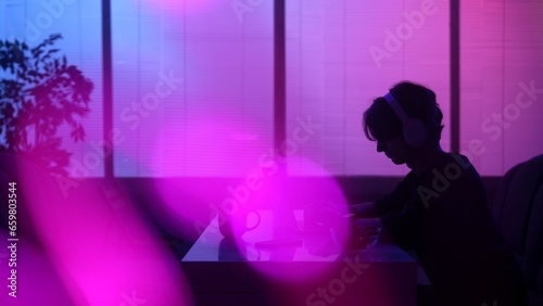 Silhouette of male sitting at the food bar at evening in neon light, wearing headphones, working on laptop.