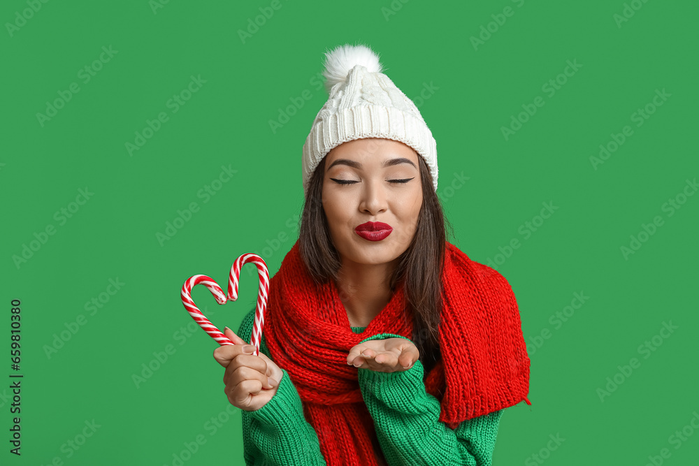 Beautiful young Asian woman in warm clothes with candy canes blowing kiss on green background Beautiful young Asian woman in warm clothes with candy canes blowing kiss on green background