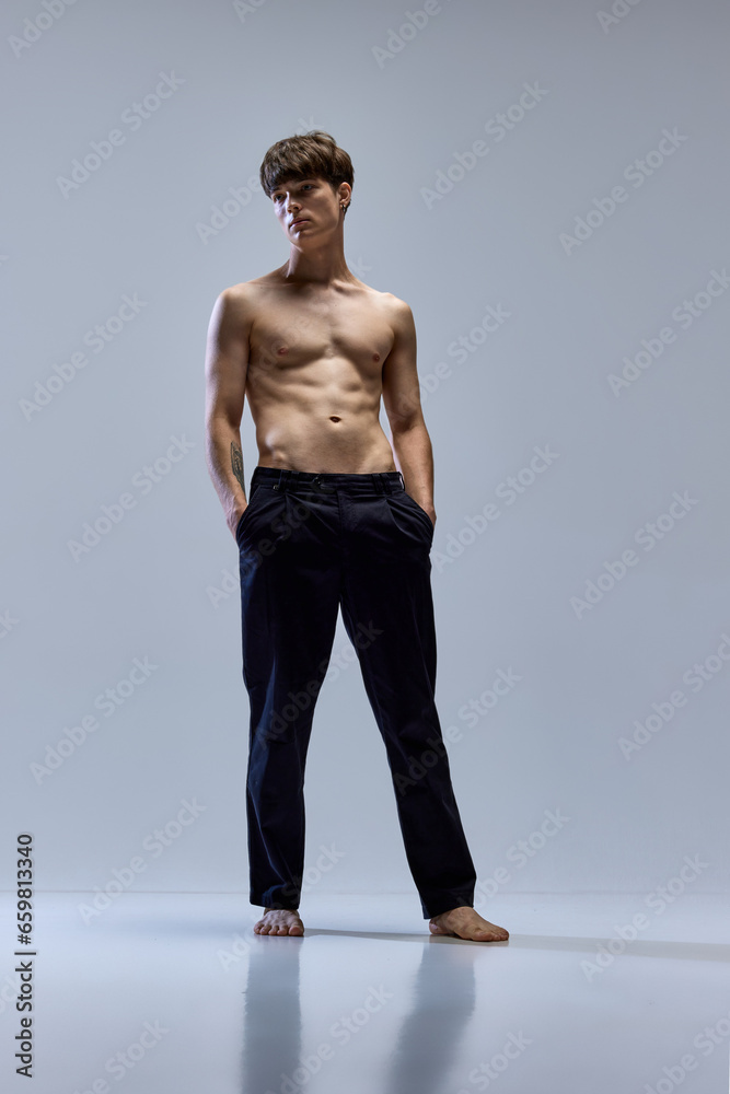 Full-length portrait of young man posing looking away in trousers isolated over grey studio background. Male muscular build body. Full-length portrait of young man posing looking away in trousers isolated over grey studio background. Male muscular build body.