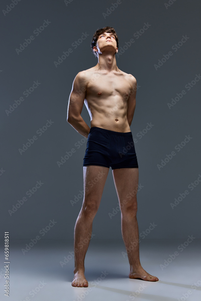 Full-length portrait of young man with naked muscular build body, torso posing against over dark grey studio background.