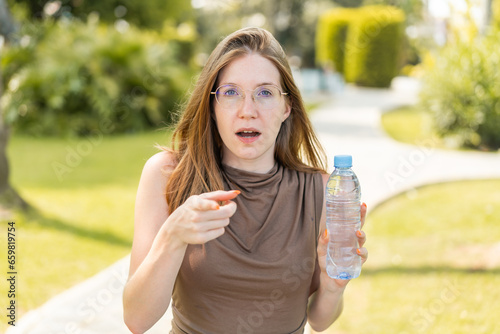 French girl with glasses holding a bottle of water at outdoors surprised and pointing front