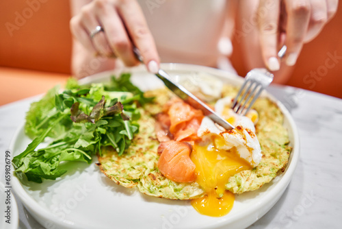 Woman cuts Zucchini pancakes  with avocado  cheese cream  salmon and egg . Healthy breakfast  protein. Restaurant dish. Breakfast in cafe on a sunny morning  on the summer veranda