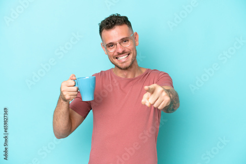 Young caucasian man holding cup of coffee isolated on blue background points finger at you with a confident expression