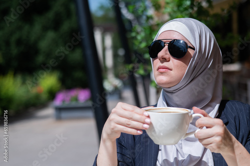 Caucasian woman in hijab drinking coffee in outdoor cafe. 