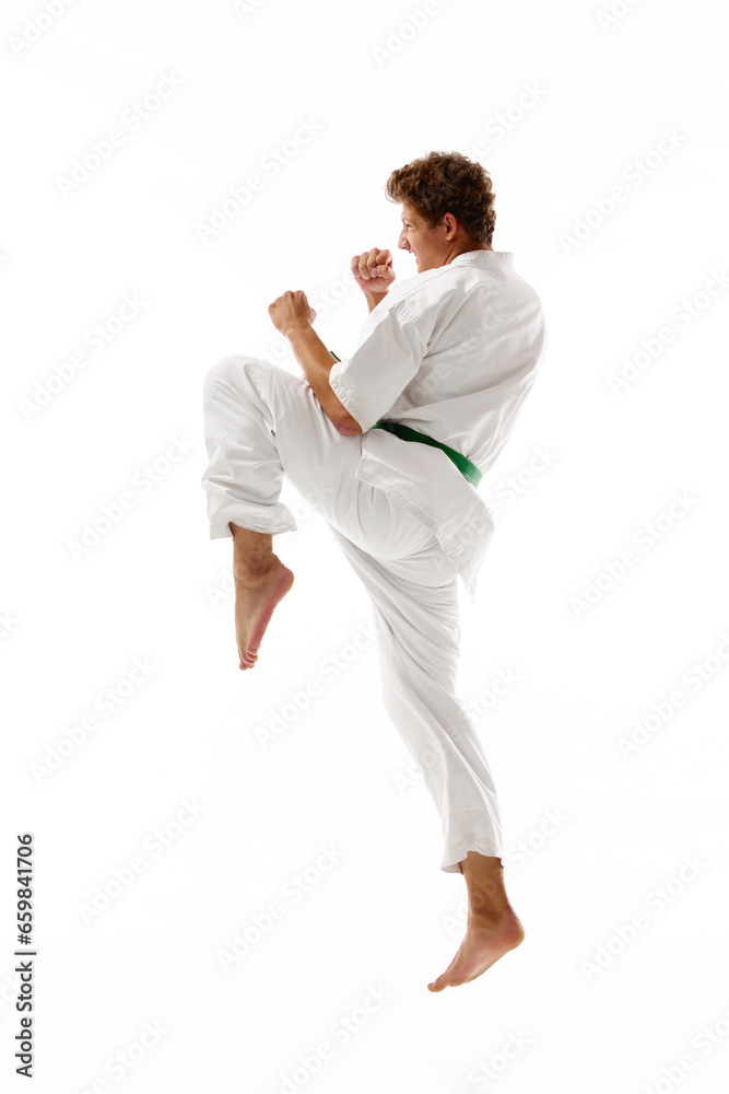 Athletic young man, karateka in white kimono and green belt training, practicing karate isolated on white studio background. Concept of martial arts, combat sport, energy, strength, health. Ad