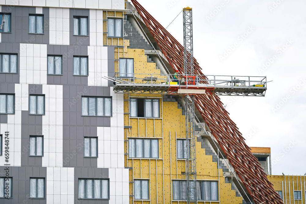 Worker insulate walls of building facade. Residential building under construction, man work in construction cradle. Exterior wall heat insulation with mineral wool. Cladding and warming wall