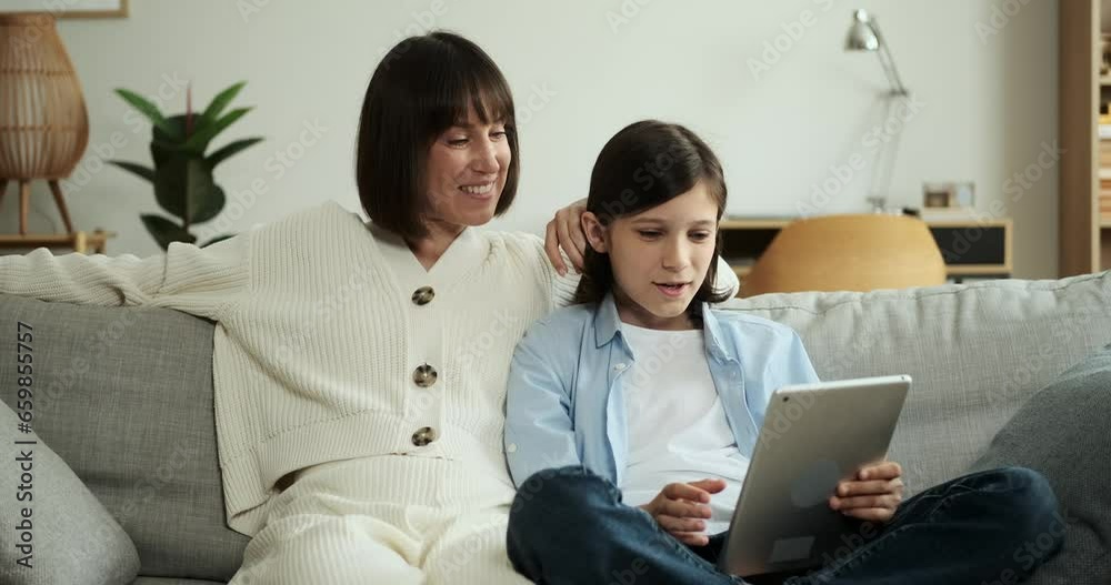 Mother and son engage in a conversation using a digital tablet while sitting on the living room couch. Their dialogue and connection create a communicative atmosphere. Mother and son engage in a conversation using a digital tablet while sitting on the living room couch. Their dialogue and connection create a communicative atmosphere.