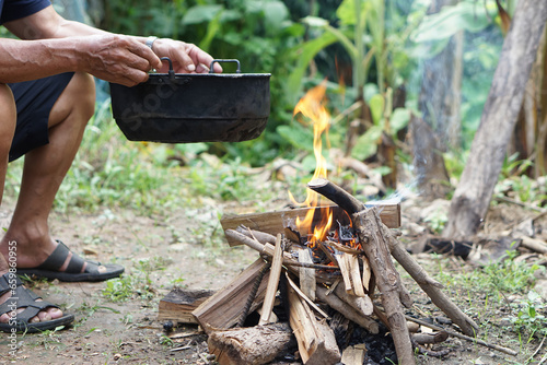 Close up man holds old black pot to cook on bonfire. Concept, cooking outdoor, kitchen in forest. Survival life skill for camping or hiking. Rural traditional lifestyle. Lighting fire with wood. photo