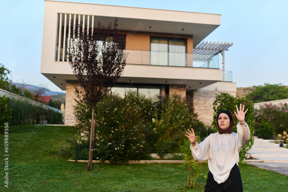 A modern hijab-clad woman stands in front of a contemporary villa, with raised hands simulating interactions with holograms and smart objects, highlighting the space for holographic additions