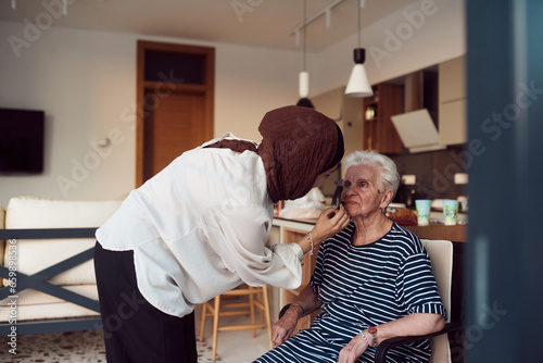 In this heartwarming real-life scene, a girl in a hijab and her sister lovingly apply makeup to their elderly grandmother, preparing her for a special family anniversary celebration, showcasing the