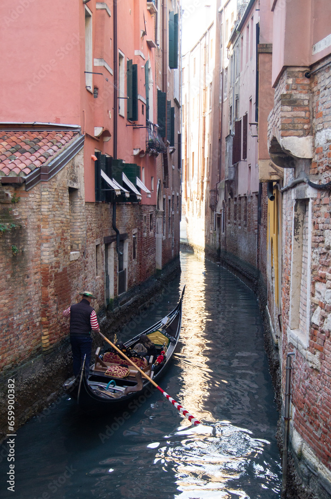 Narrow canal with one, 1 gondola filled with tourists and a gondolier. Venitian architecture, buildings, dark water in the canal, reflection on the water, evening soft light. Crumbling buildings.
