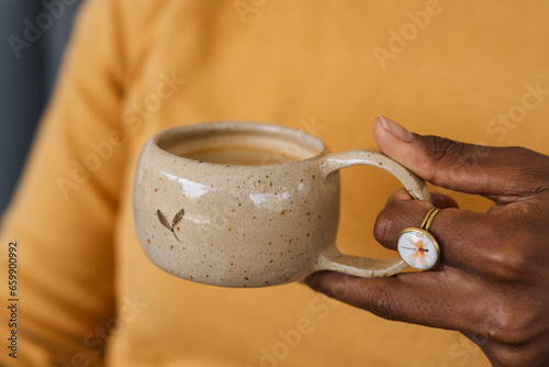 Closeup of female hands with a coffee mug of beverage. Beautiful dark skinned girl in pastel orange sweater holding cup of tea or coffee in the morning sunlight. Clay Cup with cute heart and cute ring