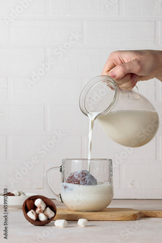 A hand pours milk from a jug into a glass cup with cocoa, chocolate bomb. Making hot chocolate. Vertical frame, light background.