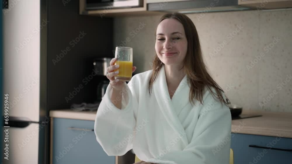 I enjoy a well-balanced breakfast. A smiling woman in a bathrobe drinks orange juice. at the kitchen table. Young beautiful woman in the kitchen drinking juice. I enjoy a well-balanced breakfast. A smiling woman in a bathrobe drinks orange juice. at the kitchen table. Young beautiful woman in the kitchen drinking juice.